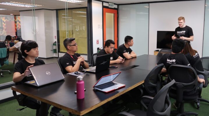 LevelUp team in a meeting room, with several members seated at a large table, attentively listening to a presenter standing at the front, with laptops and phones on the table.